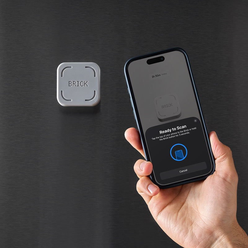 Hand holding a smartphone with app interface next to a 'BRICK' device on a dark background